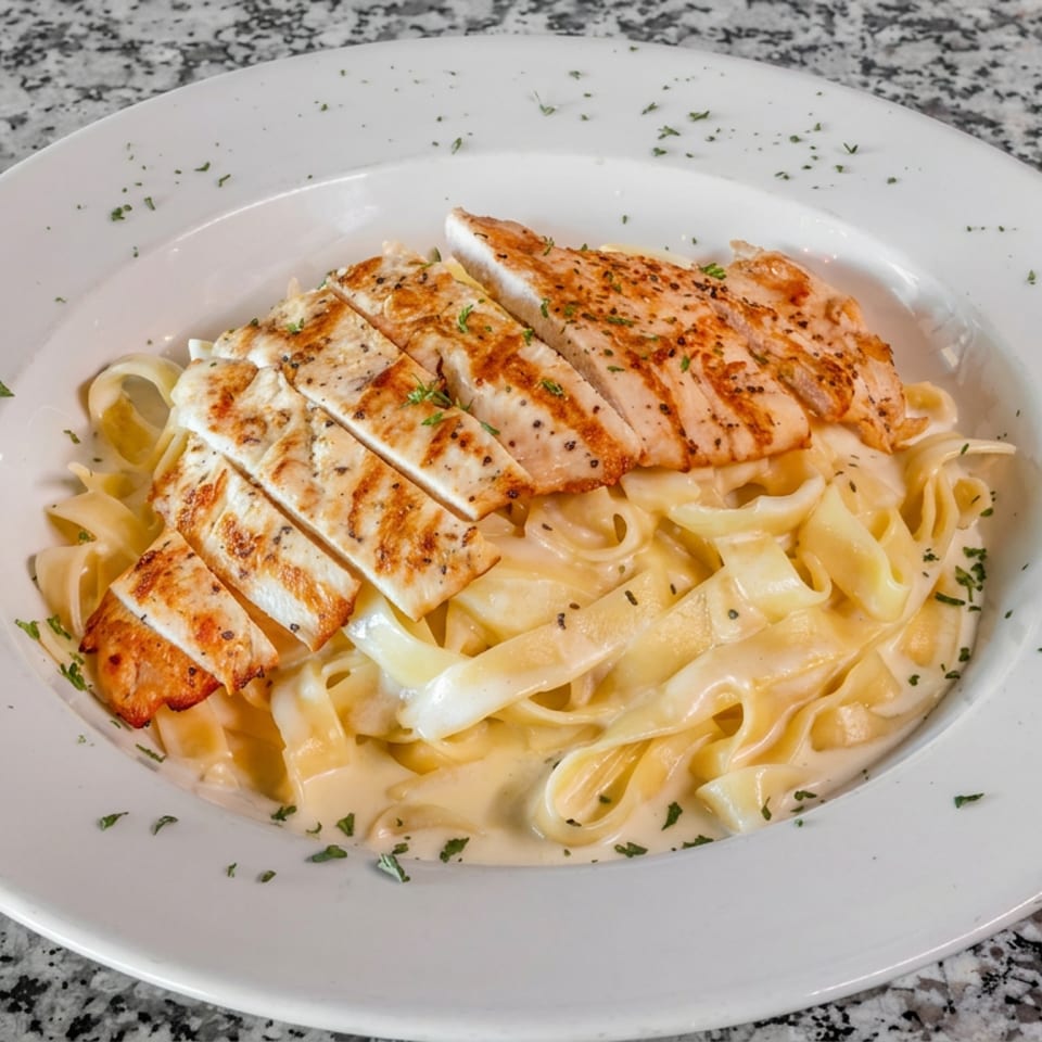 Sliced grilled chicken served over creamy fettuccine Alfredo pasta, garnished with herbs on a white plate.